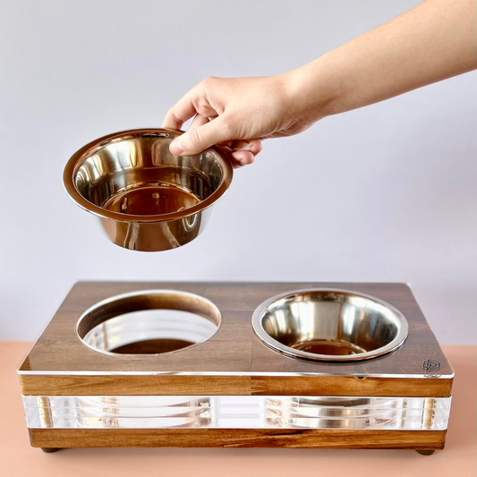 A hand holding a stainless steel bowl above a wooden pet feeder with a second bowl, featuring a sleek design with acrylic and reclaimed timber.